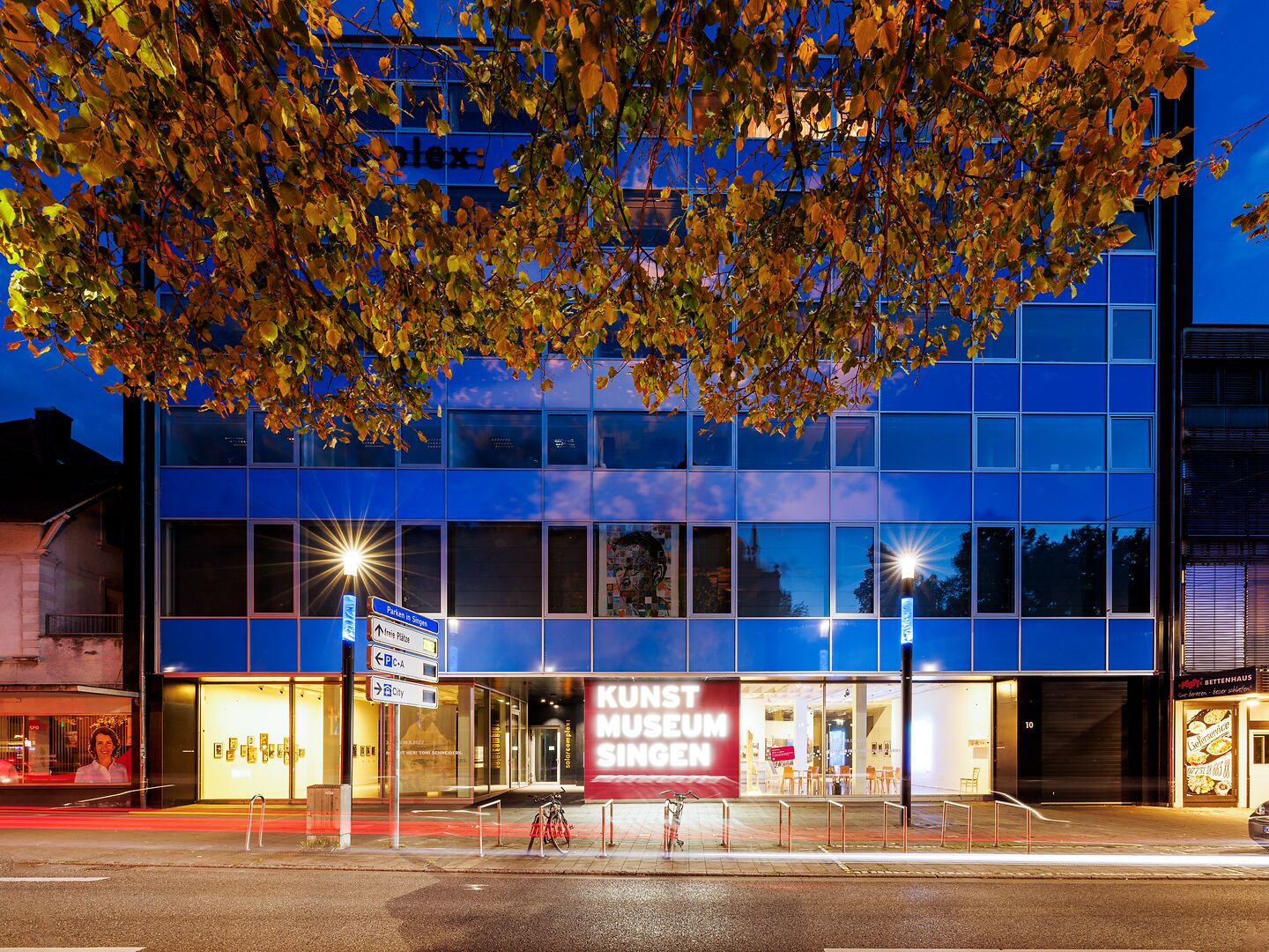 Kunstmuseum Singen bei Nacht, modernes Gebäude mit blauer Glasfassade, beleuchtet und von einem herbstlichen Baum eingerahmt